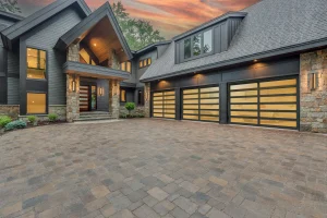A luxurious modern estate featuring a spacious paver driveway and three black-framed glass sectional doors glowing warmly at dusk, perfectly capturing the aesthetic impact of choosing a new garage door.