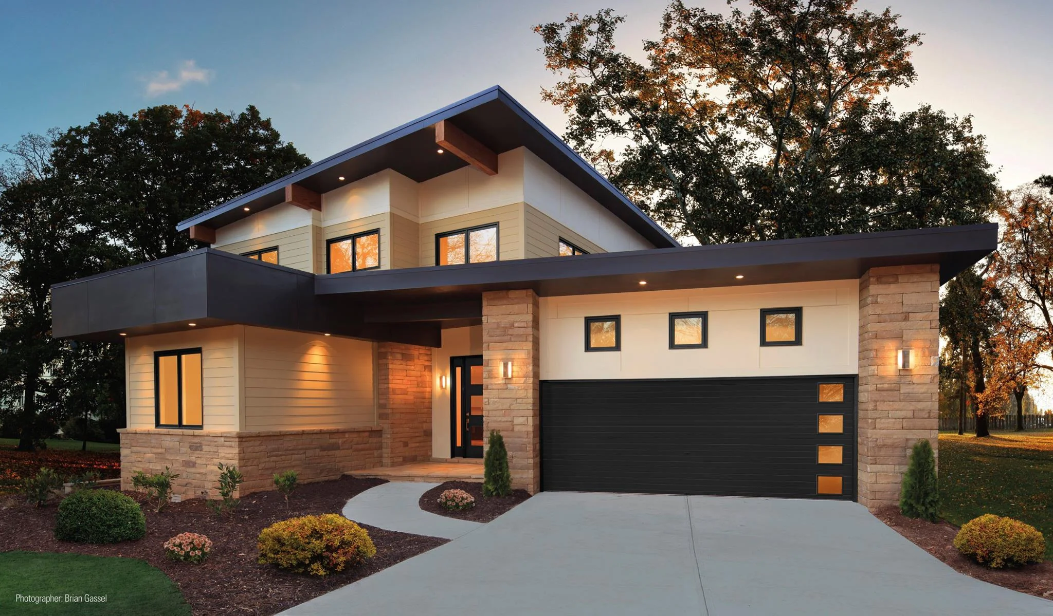 A stunning two-story contemporary home featuring stone accents, warm exterior lighting, and a matte black garage door with vertically stacked frosted square windows along the side, showcasing the visual appeal of custom garage doors.