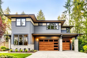 A beautiful multi-story modern home with grey siding and stone accents featuring natural wood sectional doors with asymmetrical tinted windows, emphasizing the beauty and integration of a home security garage door.