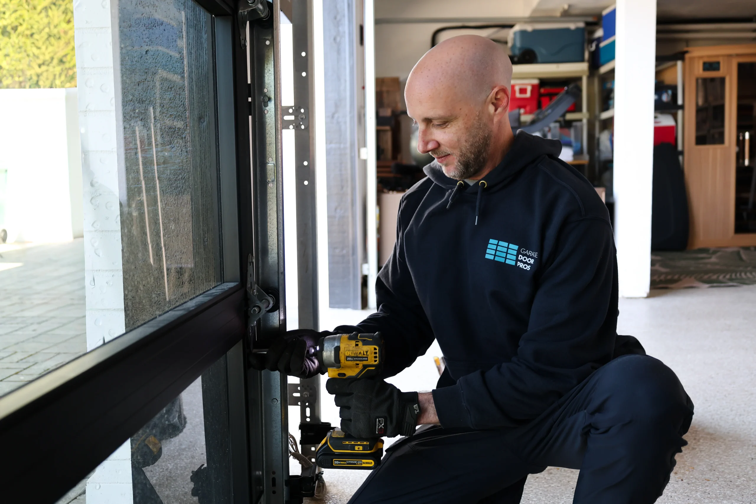 A professional technician uses an impact driver to carefully adjust and secure the side hinges on a sleek, black-framed glass sectional door during a modern garage door installation.