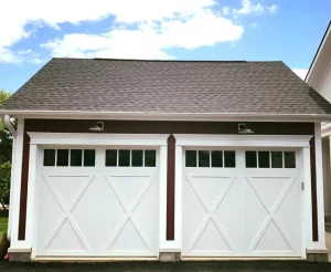 An exterior view of a detached garage with two white, carriage-house style doors featuring prominent X-shaped paneling and top windows, highlighting modern updates often integrated with smart garage door security.